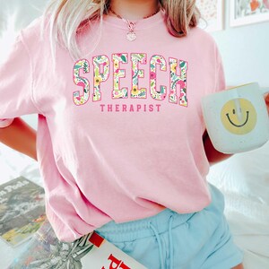 May include: Pink t-shirt with the text "SPEECH THERAPIST" in floral letters. The letters are pink with a white outline and are filled with a floral pattern.