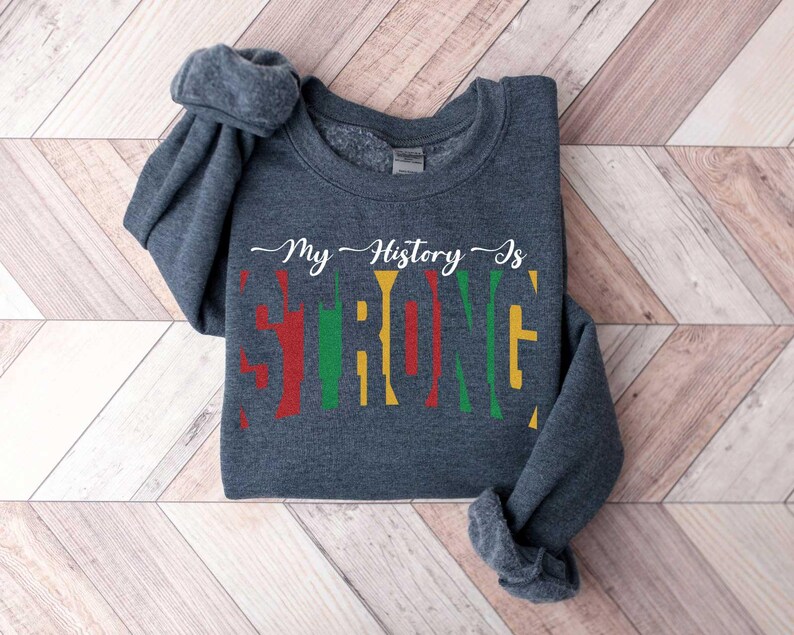 May include: A dark grey sweatshirt with the text "My History Is" and the word "Strong" in colourful block letters. The letters are in red, yellow, and green.