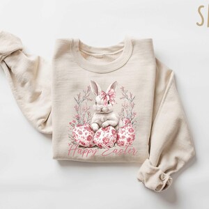 May include: A beige sweatshirt with a white bunny rabbit sitting on pink and white floral eggs. The text "Happy Easter" is printed below the bunny.