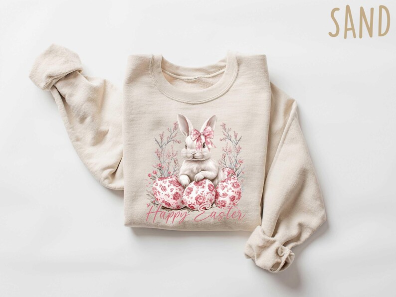 May include: A beige sweatshirt with a white bunny rabbit sitting on pink and white floral eggs. The text "Happy Easter" is printed below the bunny.