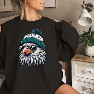 Puede incluir: Sudadera negra con un gr&aacute;fico de un &aacute;guila calva con una boina verde y blanca con la palabra "PHILADELPHIA" en ella. El &aacute;guila lleva gafas de sol.
