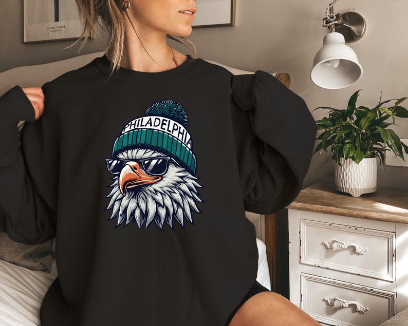 Puede incluir: Sudadera negra con un gr&aacute;fico de un &aacute;guila calva con una boina verde y blanca con la palabra "PHILADELPHIA" en ella. El &aacute;guila lleva gafas de sol.