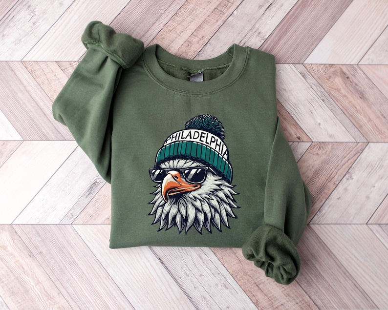 Puede incluir: Una sudadera verde con un gr&aacute;fico de un &aacute;guila con gafas de sol y una boina con el texto "PHILADELPHIA" en ella.