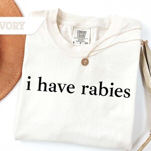 Może przedstawiać: Koszulka w kolorze kości słoniowej z nadrukiem "i have rabies" w kolorze czarnym.