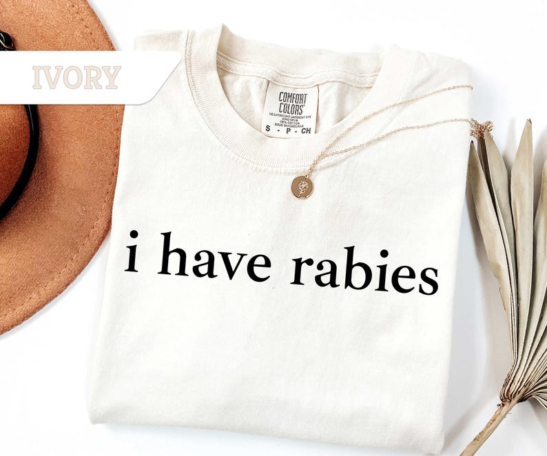 Może przedstawiać: Koszulka w kolorze kości słoniowej z nadrukiem "i have rabies" w kolorze czarnym.