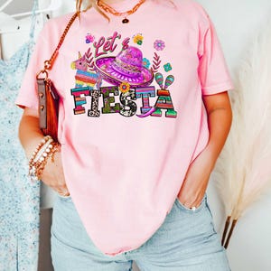 May include: Pink t-shirt with the words "Let's Fiesta" in colorful letters and a sombrero graphic. The shirt features a festive design with flowers and a pi&ntilde;ata, perfect for a celebration.