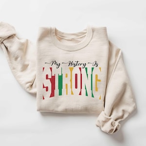 May include: A beige sweatshirt with the text "My History Is Strong" printed in black, red, yellow, and green. The text is styled to look like a stacked bar graph.