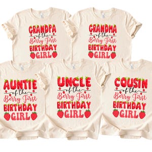 May include: Five cream-colored t-shirts with text that reads "Grandpa", "Grandma", "Auntie", "Uncle", and "Cousin" above "of the Berry First Birthday Girl" in red and pink strawberry-themed font. Each shirt has a strawberry graphic.