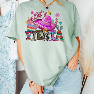 May include: Light green t-shirt with a colorful "Let's Fiesta" graphic. The design includes a sombrero, flowers, and a pi&ntilde;ata. The word "FIESTA" is in bold, multi-colored letters. The shirt is a casual, festive style.