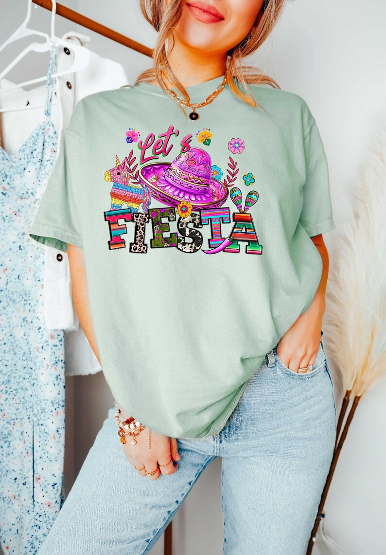 May include: Light green t-shirt with a colorful "Let's Fiesta" graphic. The design includes a sombrero, flowers, and a pi&ntilde;ata. The word "FIESTA" is in bold, multi-colored letters. The shirt is a casual, festive style.