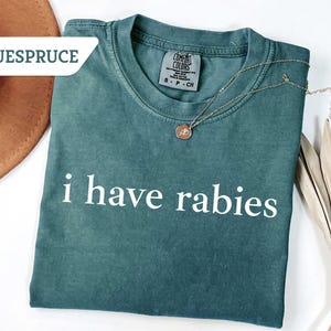 Może przedstawiać: Zielony t-shirt z nadrukiem "i have rabies" w kolorze białym na przodzie.