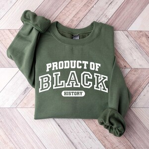 Puede incluir: Una sudadera verde con el texto "PRODUCT OF BLACK HISTORY" en blanco.