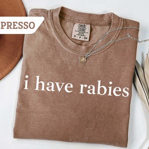 Może przedstawiać: Brązowy T-shirt z nadrukiem "i have rabies" w kolorze białym. Koszulka wykonana jest z miękkiej, wygodnej tkaniny i ma luźny kr&oacute;j.