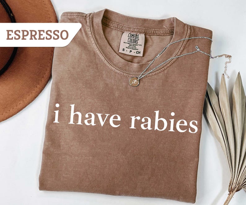 Może przedstawiać: Brązowy T-shirt z nadrukiem "i have rabies" w kolorze białym. Koszulka wykonana jest z miękkiej, wygodnej tkaniny i ma luźny kr&oacute;j.