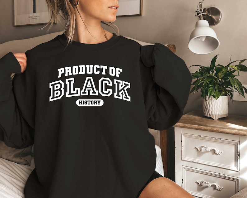 Puede incluir: Sudadera negra con texto blanco que dice "Producto de la historia negra".