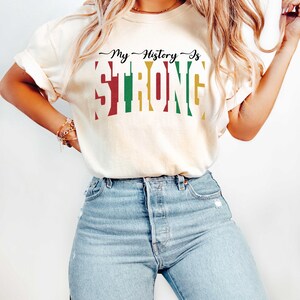 May include: A woman wearing a light beige t-shirt with the text "My History Is STRONG" in red, green, and yellow. The text is in a bold, blocky font. She is wearing light blue jeans.