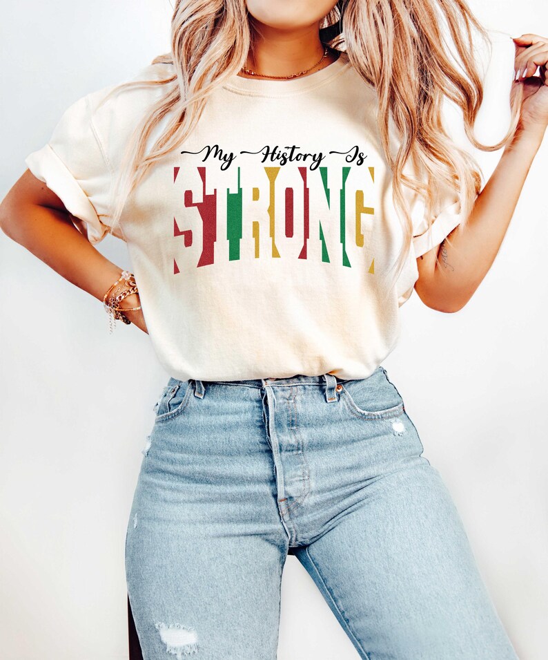 May include: A woman wearing a light beige t-shirt with the text "My History Is STRONG" in red, green, and yellow. The text is in a bold, blocky font. She is wearing light blue jeans.