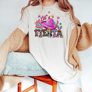 May include: Cream-colored t-shirt with a colorful "Let's Fiesta" graphic. The design includes a sombrero, flowers, and a pi&ntilde;ata. The text is in a mix of fonts and colors, creating a festive look.