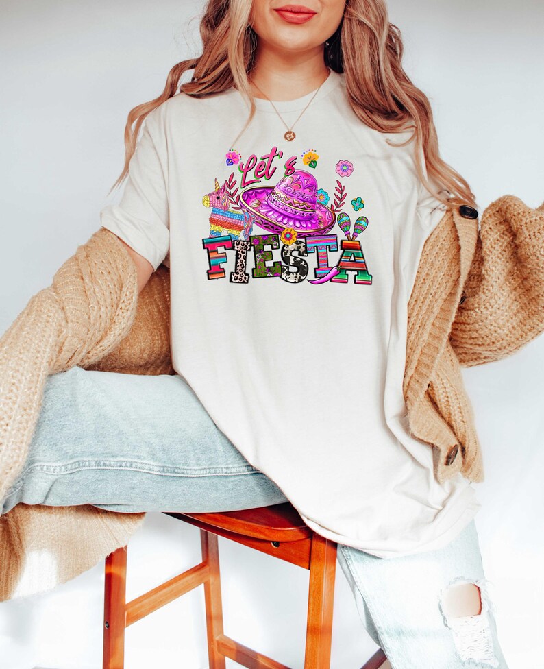 May include: Cream-colored t-shirt with a colorful "Let's Fiesta" graphic. The design includes a sombrero, flowers, and a pi&ntilde;ata. The text is in a mix of fonts and colors, creating a festive look.