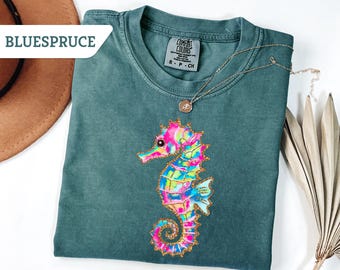 May include: A teal Comfort Colors t-shirt featuring a colorful seahorse design with pink, blue, and yellow accents. The seahorse is outlined in gold glitter. A gold necklace with a pendant is draped on the shirt. The word "BLUESPRUCE" is in the upper left corner.
