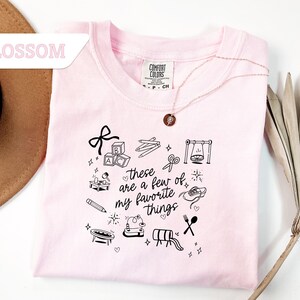 May include: A light pink t-shirt with the text "these are a few of my favorite things" and various black line art illustrations. The illustrations include a bow, blocks, pencils, scissors, a swing, a record player, a pencil, a shoe, a trampoline, a slide, and utensils.