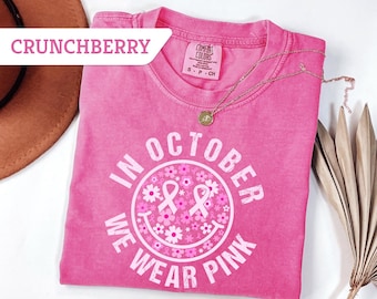 May include: A pink t-shirt with the text "IN OCTOBER WE WEAR PINK" in white, featuring a smiley face design with pink ribbons and flowers. The shirt is a Comfort Colors brand and has a small gold necklace on top.