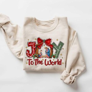 May include: A beige sweatshirt with a Christmas-themed design. The design features the word "JOY" in a stylized font with a nativity scene inside the "O". The text "To The World" is below the word "JOY".