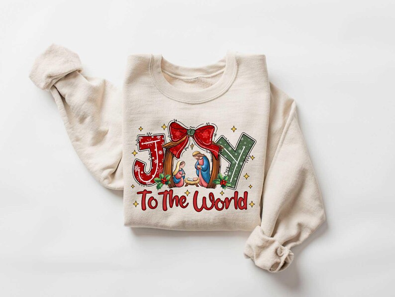 May include: A beige sweatshirt with a Christmas-themed design. The design features the word "JOY" in a stylized font with a nativity scene inside the "O". The text "To The World" is below the word "JOY".