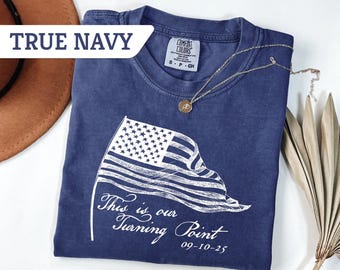 May include: A navy blue t-shirt with a white graphic of an American flag and the text "This is our Turning Point 09-10-25". The shirt is folded and has a small gold necklace on top. A brown hat and dried plant are in the background.