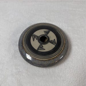 May include: A round, ceramic pottery lid with a central hole. The lid is primarily gray with a black and cream-colored circular design. The design features geometric shapes and lines. The lid is likely for a decorative jar or container.