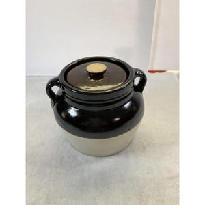 May include: A vintage stoneware crock with a lid. The crock is black on top, transitioning to a cream color at the base. It has two handles and a brown lid with a small, round knob. This piece is suitable for kitchen decor.