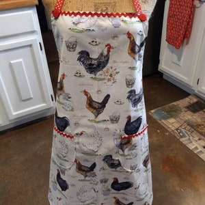 May include: A white apron with a red and white polka dot trim and a red button. The apron features a pattern of chickens, roosters, and eggs. The apron has two pockets.
