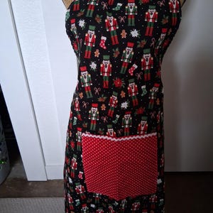 May include: A black apron with a red and white polka dot pocket. The apron is decorated with a pattern of nutcrackers, gingerbread men, and Christmas ornaments. The apron has red straps and a white lace trim on the pocket.