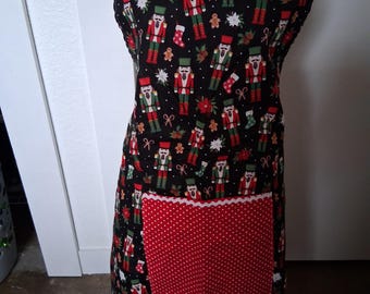 May include: A black apron with a red and white polka dot pocket. The apron is decorated with a pattern of nutcrackers, gingerbread men, and Christmas ornaments. The apron has red straps and a white lace trim on the pocket.