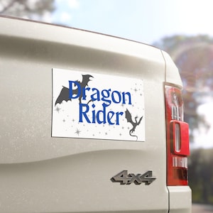 Könnte beinhalten: Ein rechteckiger weißer Autoaufkleber mit den Worten "Dragon Rider" in Blau und Schwarz, mit Drachensilhouetten und Sternen. Der Aufkleber ist an der Rückseite eines hellfarbigen Fahrzeugs angebracht.