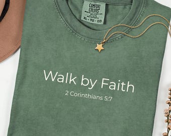 May include: A sage green t-shirt with the text "Walk by Faith" and "2 Corinthians 5:7" printed in white. A gold star pendant necklace rests on the shirt. The shirt has a Comfort Colors tag.