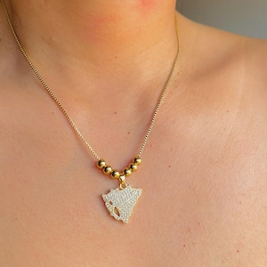 May include: A gold-colored necklace with a delicate chain. The pendant is a triangular shape, encrusted with small, sparkling crystals. The necklace features a series of gold-colored beads near the pendant, adding a touch of elegance to the jewelry.