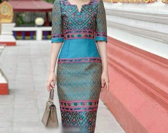 May include: A woman wearing a teal blue dress with a geometric pattern. The dress has a fitted bodice and a flowing skirt. She is carrying a brown handbag and is standing in front of a red and gold building.