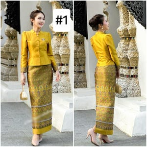 May include: A woman is wearing a yellow long-sleeved blouse with a high collar and a matching skirt with a gold and brown patterned design. The skirt is long and falls to her ankles. She is standing in front of a stone wall with a number "#1" written on it.