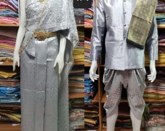 May include: A silver traditional Thai outfit with a long skirt and a shawl. The outfit is decorated with intricate patterns and embellishments. The outfit is worn by a person standing in front of a shelf of colorful fabrics. The text #1 is visible in the image.