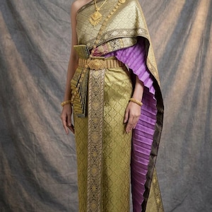 Puede incluir: Un vestido tradicional tailandés con un corpiño y falda de estampado dorado, junto con una banda morada plisada sobre un hombro. El conjunto incluye un cinturón dorado, pendientes dorados y un collar, con el cabello recogido con horquillas.