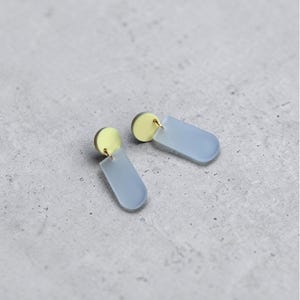 Può includere: Un paio di orecchini moderni. Ogni orecchino presenta un piccolo perno rotondo color oro collegato a un pendente rettangolare azzurro con bordi arrotondati. Gli orecchini sono esposti su una superficie grigia.