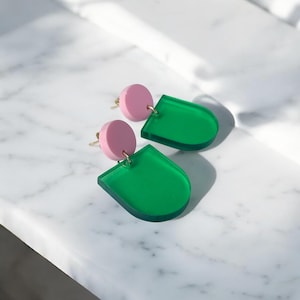 Könnte beinhalten: Ein Paar moderne Ohrringe mit einem rosa kreisförmigen Ohrstecker und einer grünen, halbtransparenten, geometrischen Form. Die Ohrringe sind mit einem goldfarbenen Draht verbunden. Die Ohrringe werden auf einer weißen Marmoroberfläche präsentiert.
