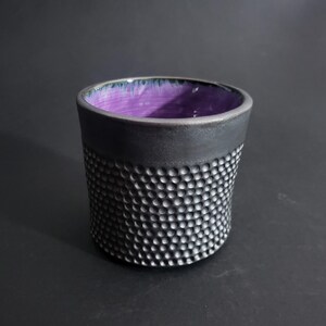 May include: A handcrafted ceramic cup with a textured, dimpled exterior and a dark gray rim. The interior is a vibrant purple, creating a striking contrast. This unique cup is perfect for coffee, tea, or other beverages.