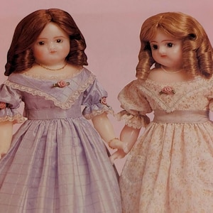May include: Two antique porcelain dolls with auburn hair and delicate features. They wear long dresses, one lavender with a plaid pattern, the other a floral print. Both dolls have rose accents and hold hands, showcasing vintage charm.