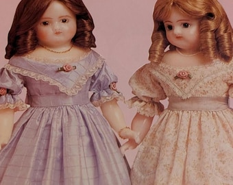 Op de afbeelding: Twee antieke porseleinen poppen met rossig haar en delicate trekken. Ze dragen lange jurken, één lavendelkleurig met een ruitpatroon, de andere met een bloemenprint. Beide poppen hebben rozenaccenten en houden elkaars hand vast, wat de vintage charme laat zien.