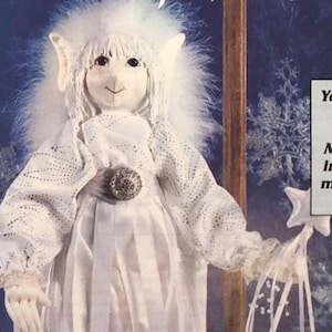 Peut inclure: Poupée artisanale de Jack Frost, vêtue d'une tenue blanche, comprenant une robe, des manches et des bordures à volants. La poupée a un visage souriant, des cheveux blancs et une baguette en forme d'étoile. Le texte "Jack Frost" est en haut.