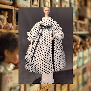 Könnte beinhalten: Eine Vintage-Puppe in einem weißen Kleid mit schwarzen Punkten. Das Kleid hat lange Ärmel mit Spitzenbesatz und einen schwarzen Schleifengürtel. Die Puppe hat ein bemaltes Gesicht und steht auf einer weißen Basis.