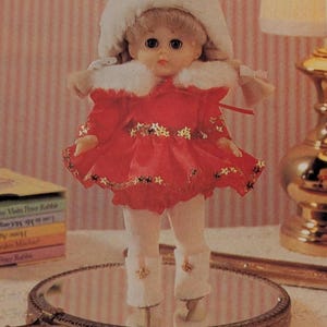 May include: A vintage doll wearing a red dress with gold star accents, a white fur-trimmed collar, and a matching hat. The doll has blonde hair and is wearing white pants and boots. The doll is standing on a mirrored surface.