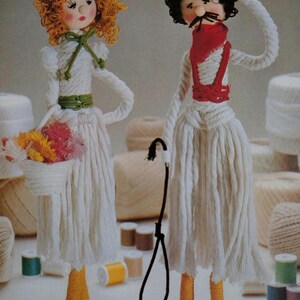 May include: Handmade yarn dolls, a woman with orange yarn hair and a man with a mustache and hat. The woman doll holds a basket of flowers. The man doll has a red scarf and suspenders. Both dolls are wearing white dresses and have yellow and white feet.